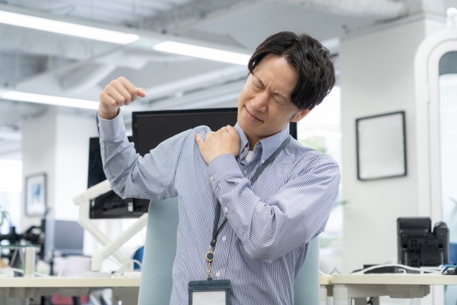 オフィスで片腕を上げ、肩に手を当てて顔をしかめる男性
