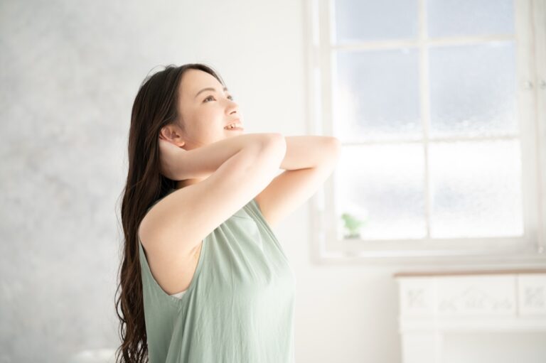 窓辺で両腕を首の後ろに回し、上を向いて笑う女性