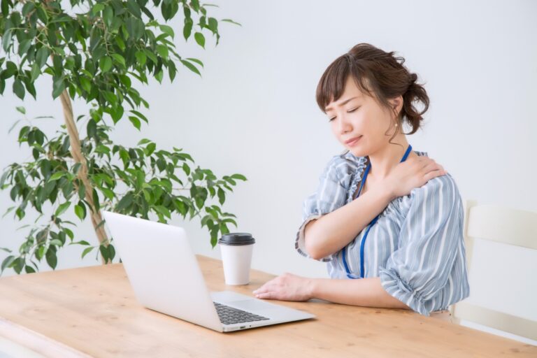 ノートパソコンの前で肩を押さえて顔をしかめる女性