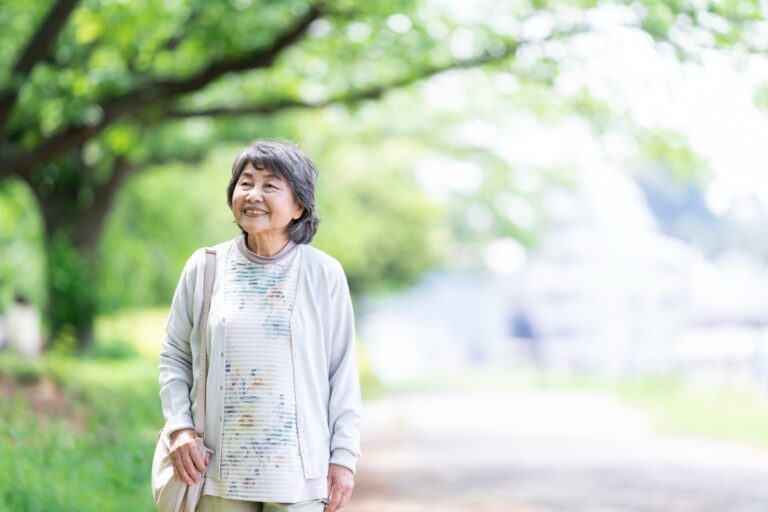 新緑の並木道で、肩にかばんをかけて微笑む年配の女性
